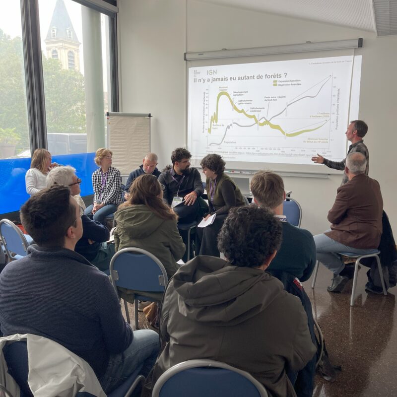 5 intervenants et 1 animateur pour une table ronde. L'intervenant montre un graphique qui est projeté au mur.