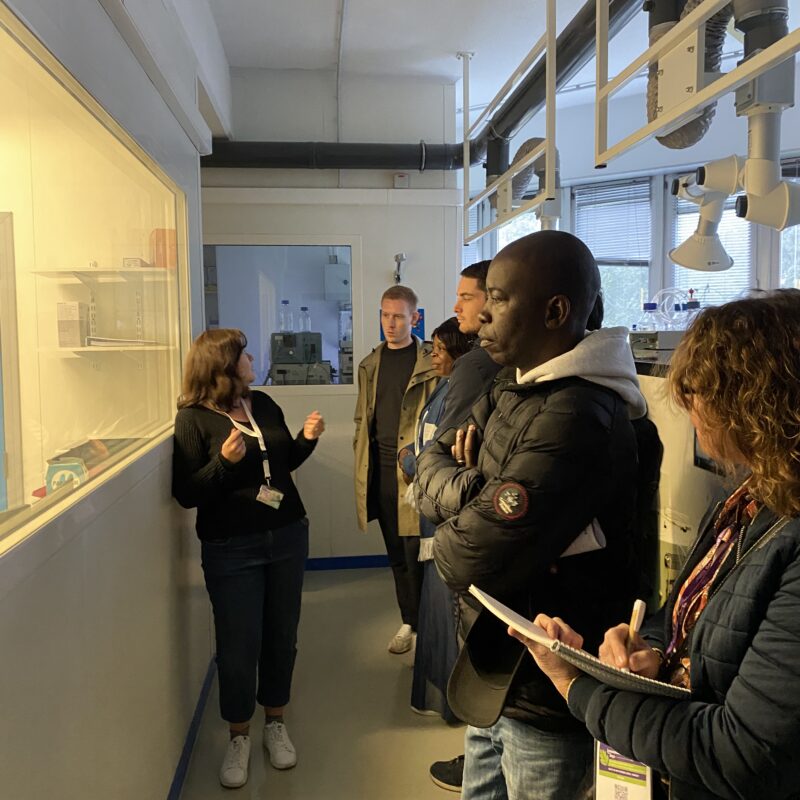 Visite du laboratoire LIBio présentée par une chercheuse : les invités prennent des notes et regardent dans une salle d'expérimentation à travers une vitre