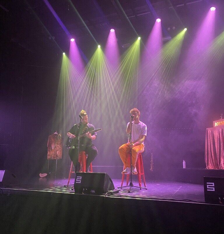 Photo des 2 chanteurs du concert de clôture à l'autre canal. Ils sont sur scène et chantent dans une atmosphère rose/violette. L'un deux a une guitare en plus